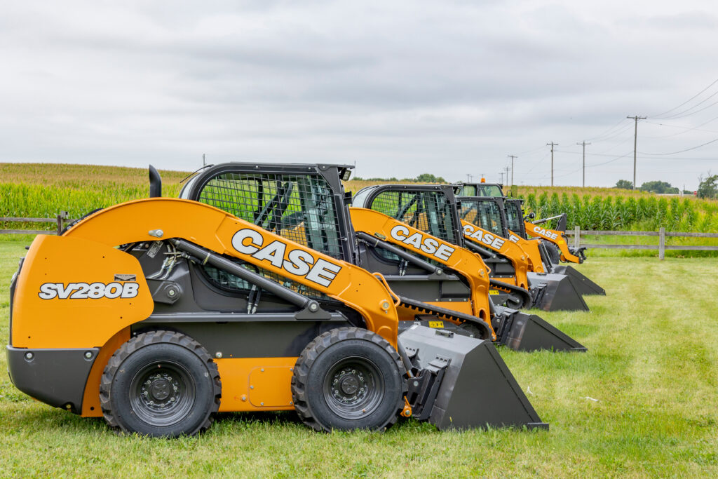 A row of SV208B Case skid loaders in a grass field with a corn field, electric poles and a cloudy sky in the background.