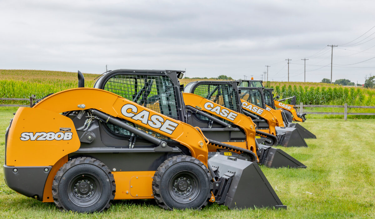 A row of SV208B Case skid loaders in a grass field with a corn field, electric poles and a cloudy sky in the background.