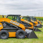 A row of SV208B Case skid loaders in a grass field with a corn field, electric poles and a cloudy sky in the background.