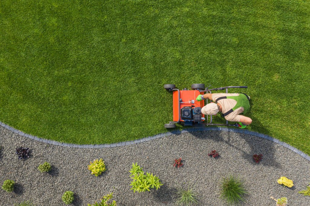 Top-down view of a landscaper mowing a green lawn with a red push mower next to a curved gravel garden bed with small plants