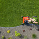 Top-down view of a landscaper mowing a green lawn with a red push mower next to a curved gravel garden bed with small plants