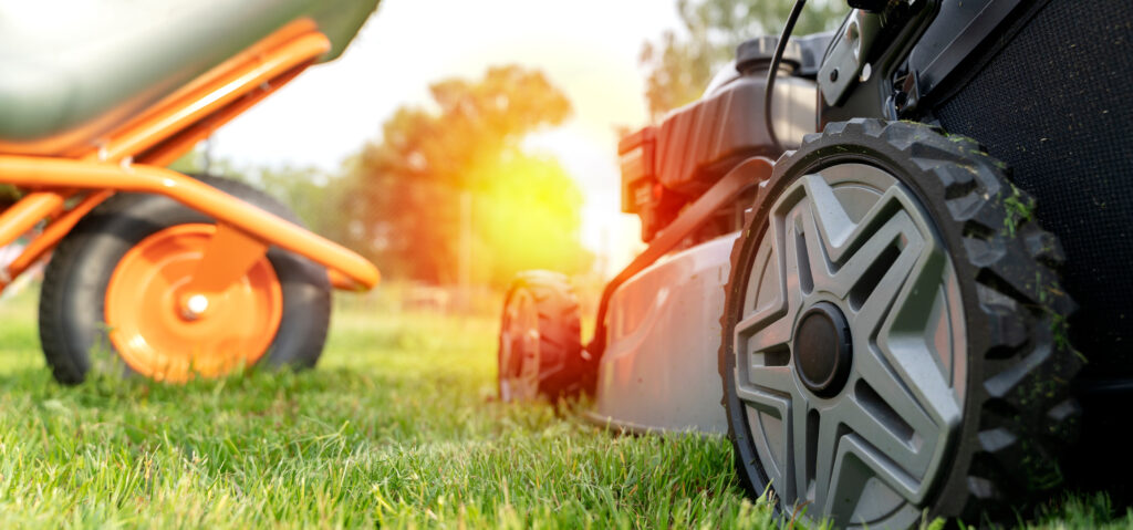 Close-up view of a lawnmower and an orange wheelbarrow on freshly cut grass with warm sunlight in the background.
