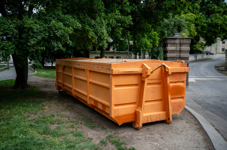 Large orange industrial dumpster placed under trees in urban park area outdoors