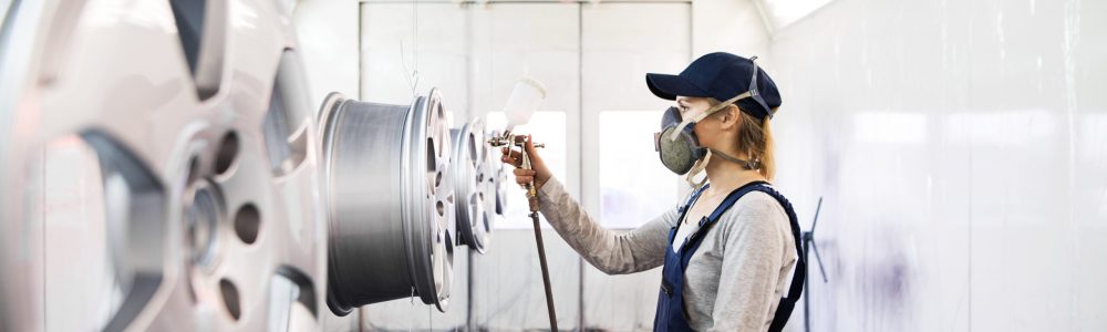 Female car painter, paingting vehicles in auto body shop. Young woman holding spray gun, spraying paint on rim of wheel, wearing mask and coveralls.